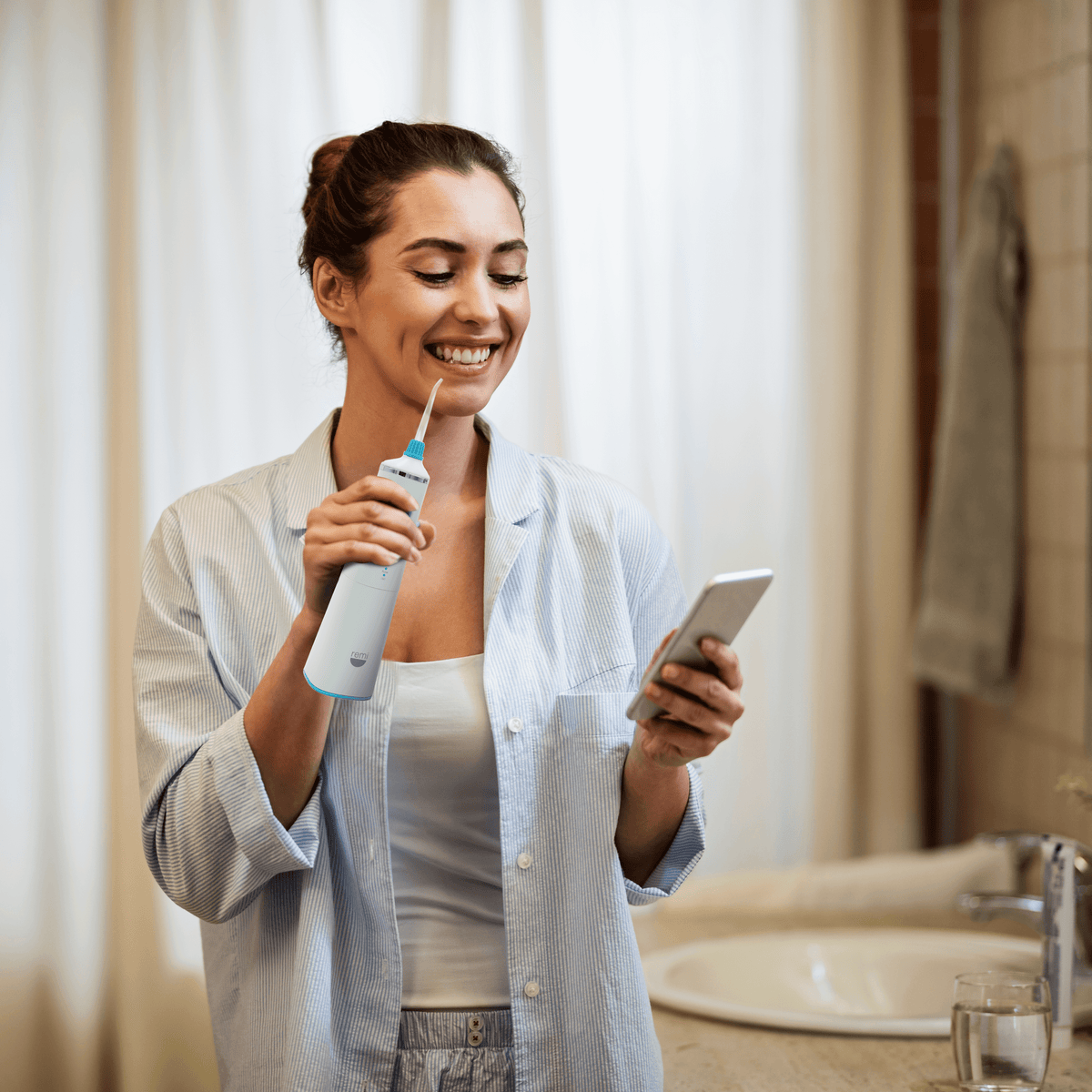 A woman smiles while holding a Waterpik Water Flosser and looking at her smartphone in the bathroom.