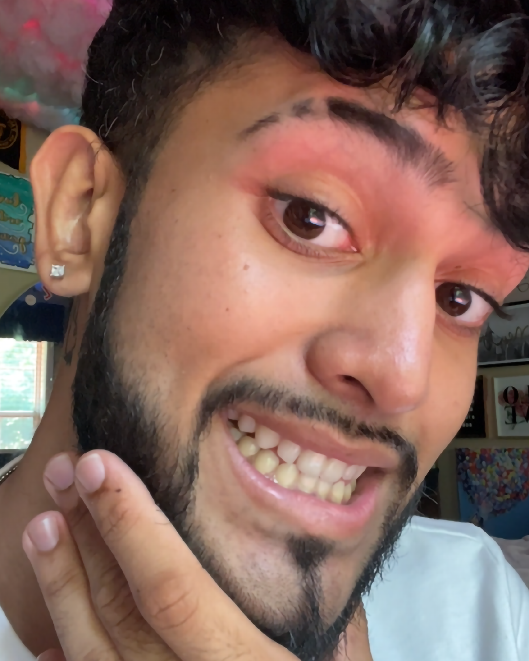 Man with a beard smiling at the camera, showing off pink eyeshadow.
