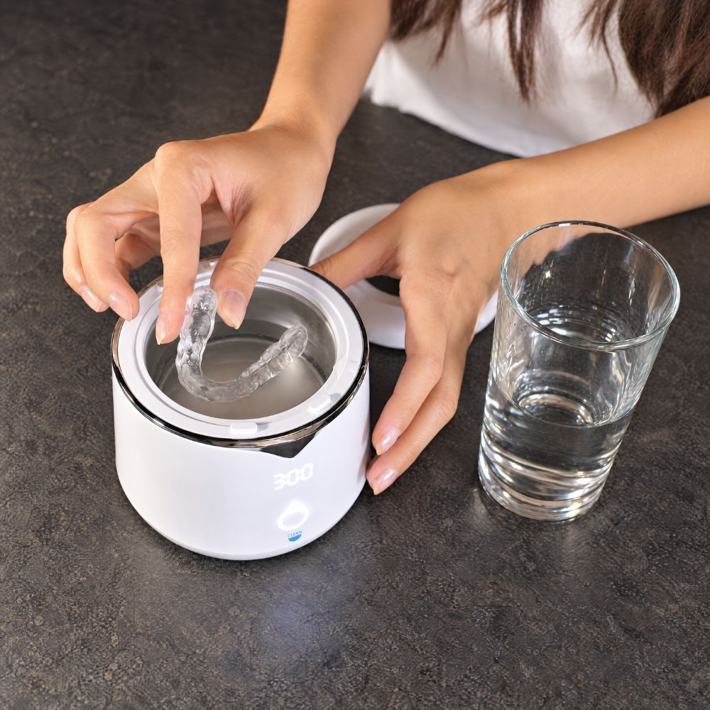 A person places a clear dental aligner into a white night guard cleaning device on a dark countertop, with a glass of water nearby.
