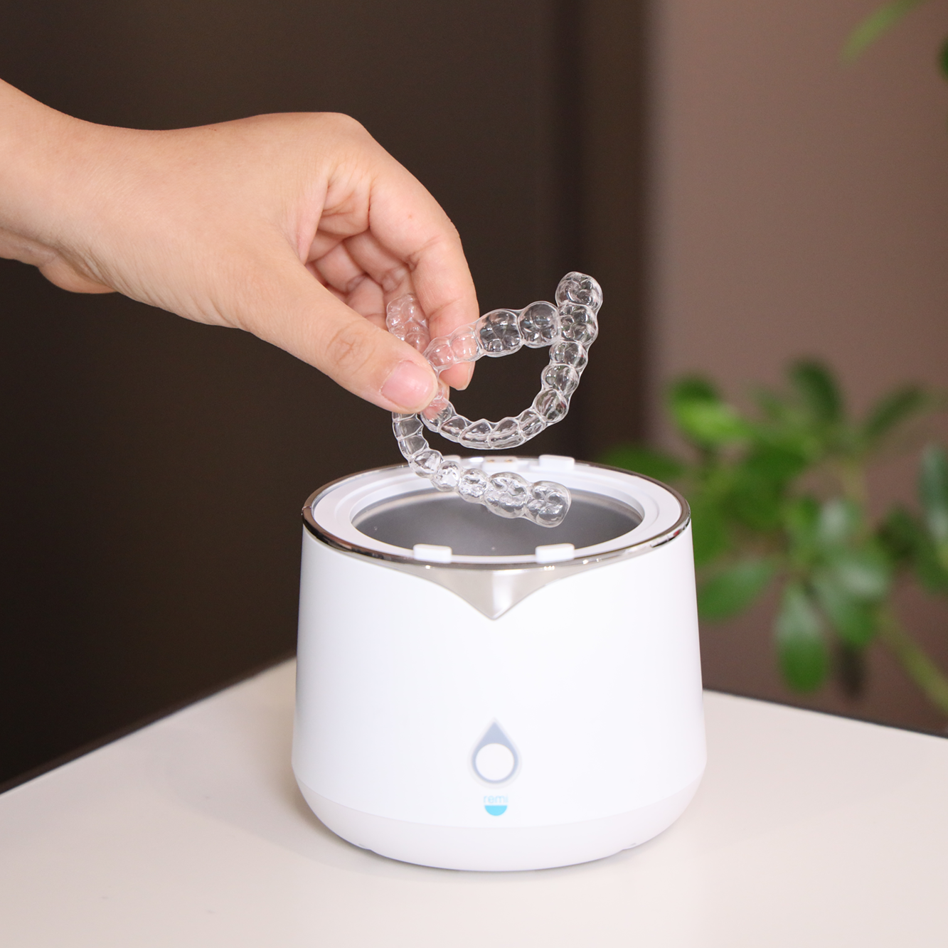 A hand is placing a clear aligner or retainer into a white ultrasonic cleaner on a table, with a green plant in the blurred background.