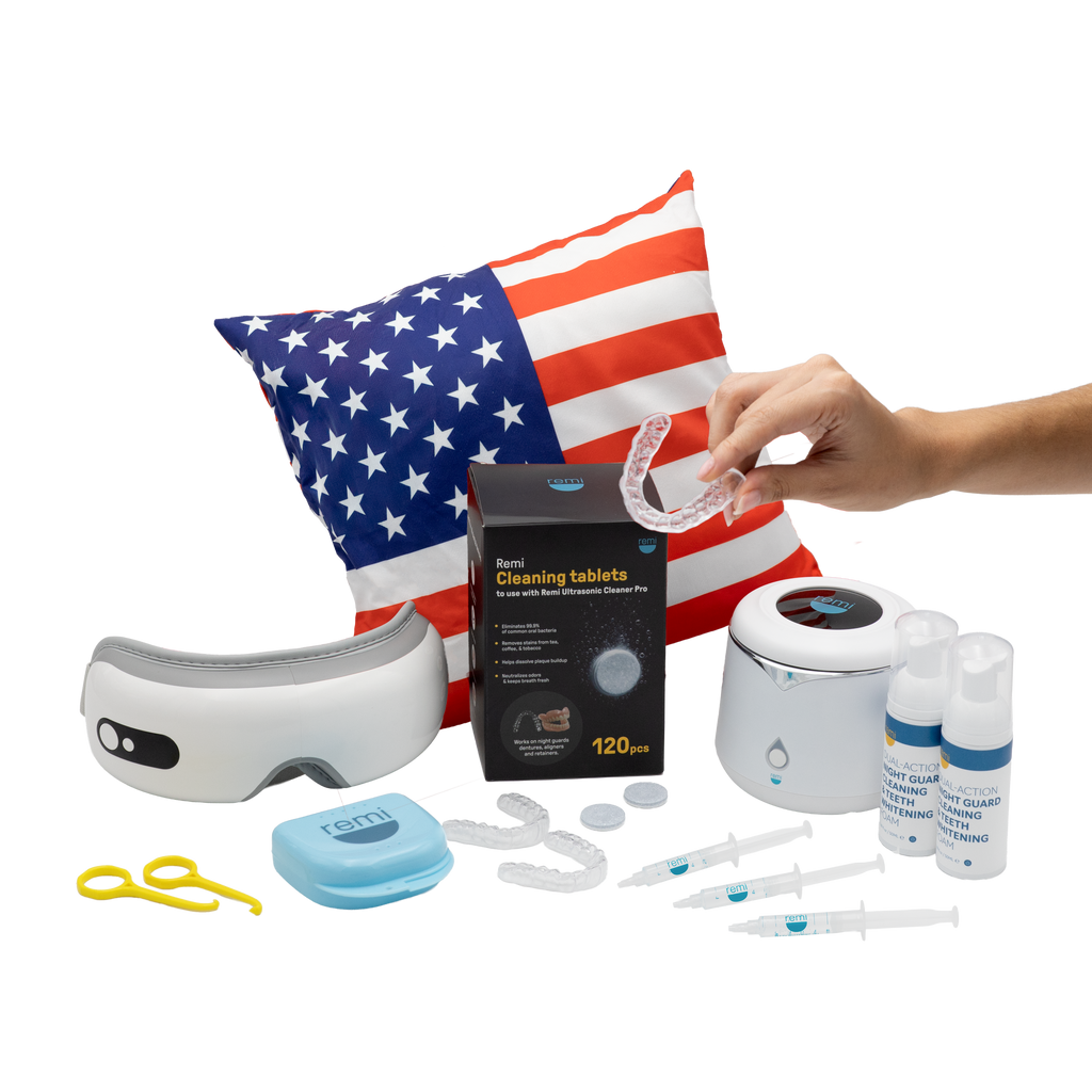 A hand holds a clear dental aligner above dental care products, including an ultrasonic cleaner, aligner cleaner, syringes, teeth whitening supplies, a UV sanitizer, a retainer case, tools, and a pillow with an American flag design.