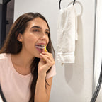 A woman with long brown hair uses a dental appliance remover while brushing her teeth and looking into the bathroom mirror. A white towel hangs on a ring beside her.