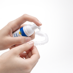 A person dispenses clear foam tray cleaning solution from a white and blue bottle onto a transparent dental retainer, holding both items against a plain white background.