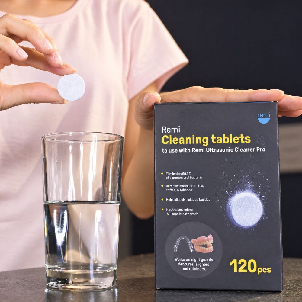 A person holds a white tablet above a glass of water next to a Remi Cleaning Tablets box for ultrasonic cleaners—ideal for custom night guard and dentist-quality oral care. Box contains 120 tablets and lists cleaning benefits.