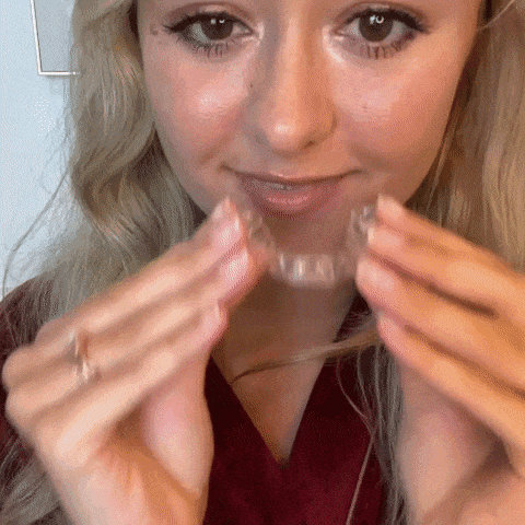 A woman is holding and adjusting a clear aligner near her mouth.