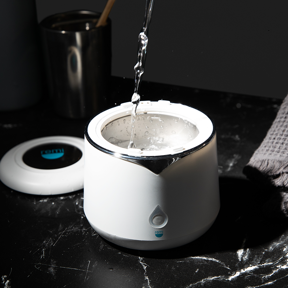 A stream of water is poured into a white electric kettle on a black marble countertop, with a lid and a textured gray towel nearby.