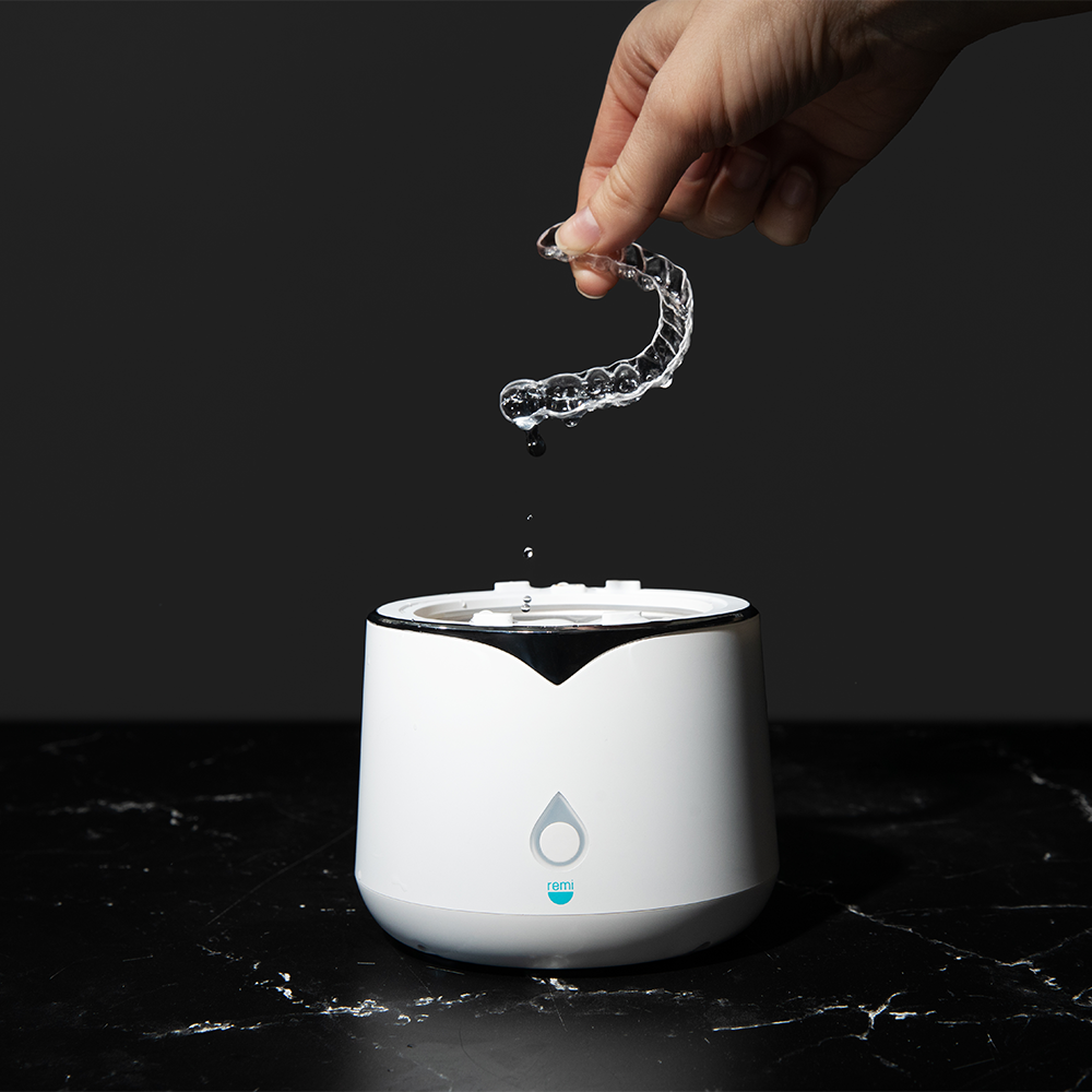 A hand holds a clear aligner above a white ultrasonic cleaner on a dark surface, with water droplets falling from the aligner into the device.