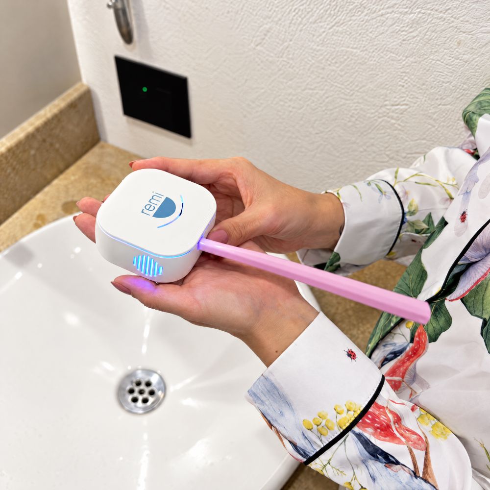 A person holds a white, square-shaped device labeled remi that emits blue light onto a pink toothbrush for plaque removal, above a bathroom sink.