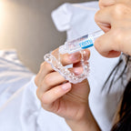 A person applies gel from a tube labeled remi onto custom night guards, holding it with both hands near a bed with white sheets.