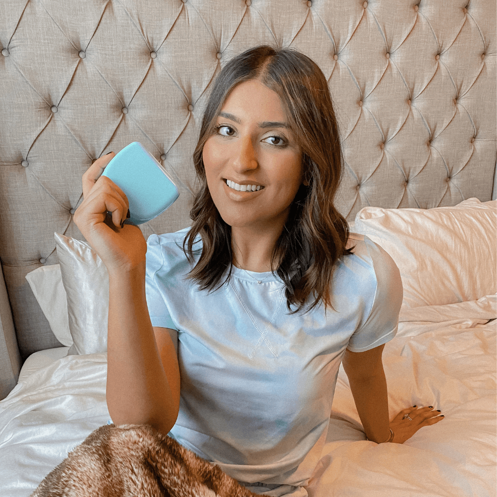 A woman with shoulder-length hair and a blue tie-dye shirt is sitting on a bed, smiling, and holding a blue square object. The bed has a tufted headboard, and there is a blanket partially visible. She seems content after using her Night Guard Cleaning + Teeth Whitening Foam (Pack of 2) before bed.