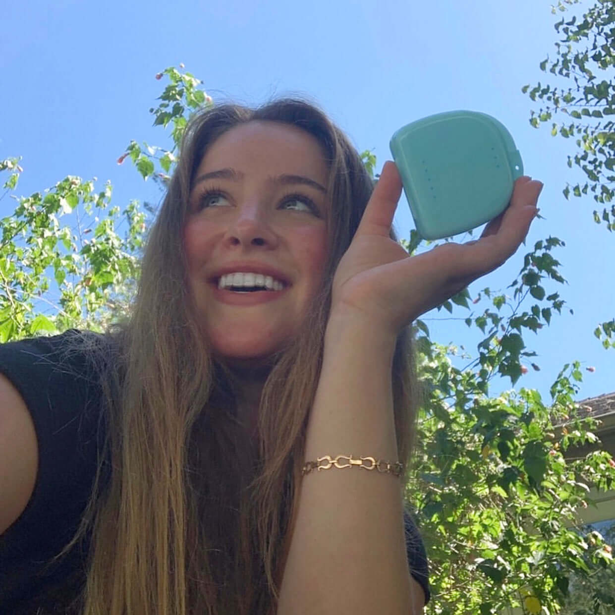 A smiling woman with long hair holds up a small, square, blue object outdoors with greenery in the background, showcasing her bright smile thanks to Night Guard Cleaning + Teeth Whitening Foam (Pack of 2).